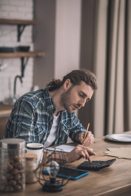 A person sitting at a desk, writing in a notebook and using a calculator. They are surrounded by papers, possibly bills or financial statements. The lighting is soft, suggesting a focused and thoughtful atmosphere.
