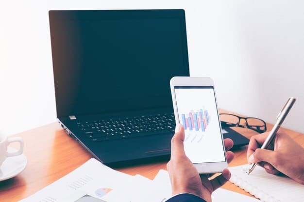 An image showing a person using a budgeting app on their smartphone, with graphs and charts displayed on the screen. The background shows a laptop with tax documents open.